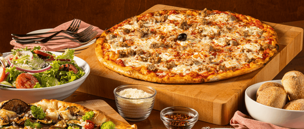 A spread of Bertucci's food with a sporkie pizza, roasted vegetable pizza, insalata salad, and rolls with crushed red pepper and parmesan cheese