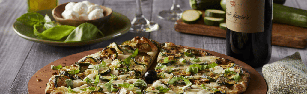 A platter from Bertucci's featuring a roasted vegetable pizza topped with fresh basil and a bottle of wine, sliced zucchini, basil, and mozzarella balls