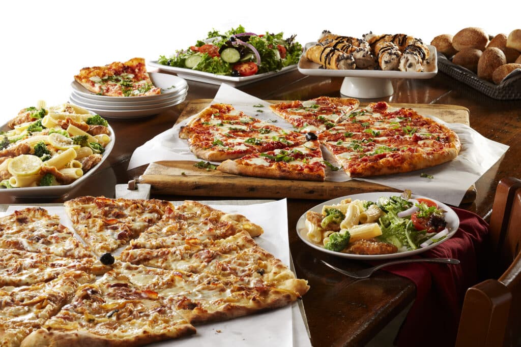 A spread of Bertucci's catering dishes featuring a margherita pizza, Rigatoni, Broccoli & Chicken pasta, Insalata salad, bread, cannoli