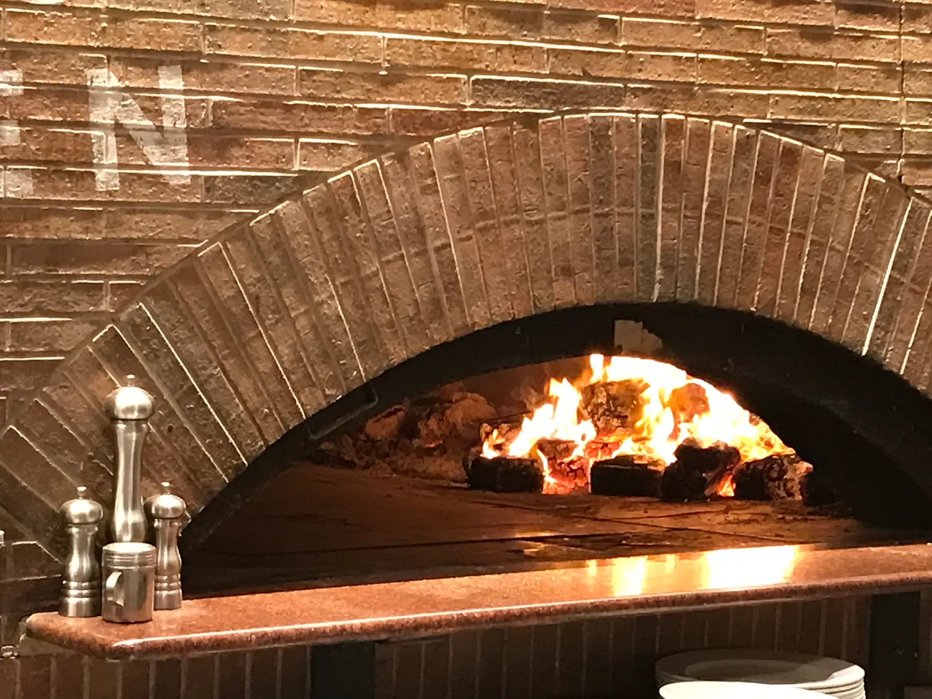 Brick pizza oven with arched brick opening and visible wood fire burning inside, set against a brick wall, with metal salt and pepper shakers on the countertop in front.