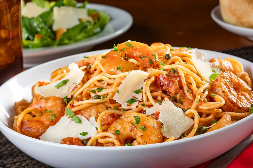 Shrimp Rossini with sautéed shrimp, spaghetti, tomatoes, capers, fresh herbs, and shaved Parmesan in a white bowl.
