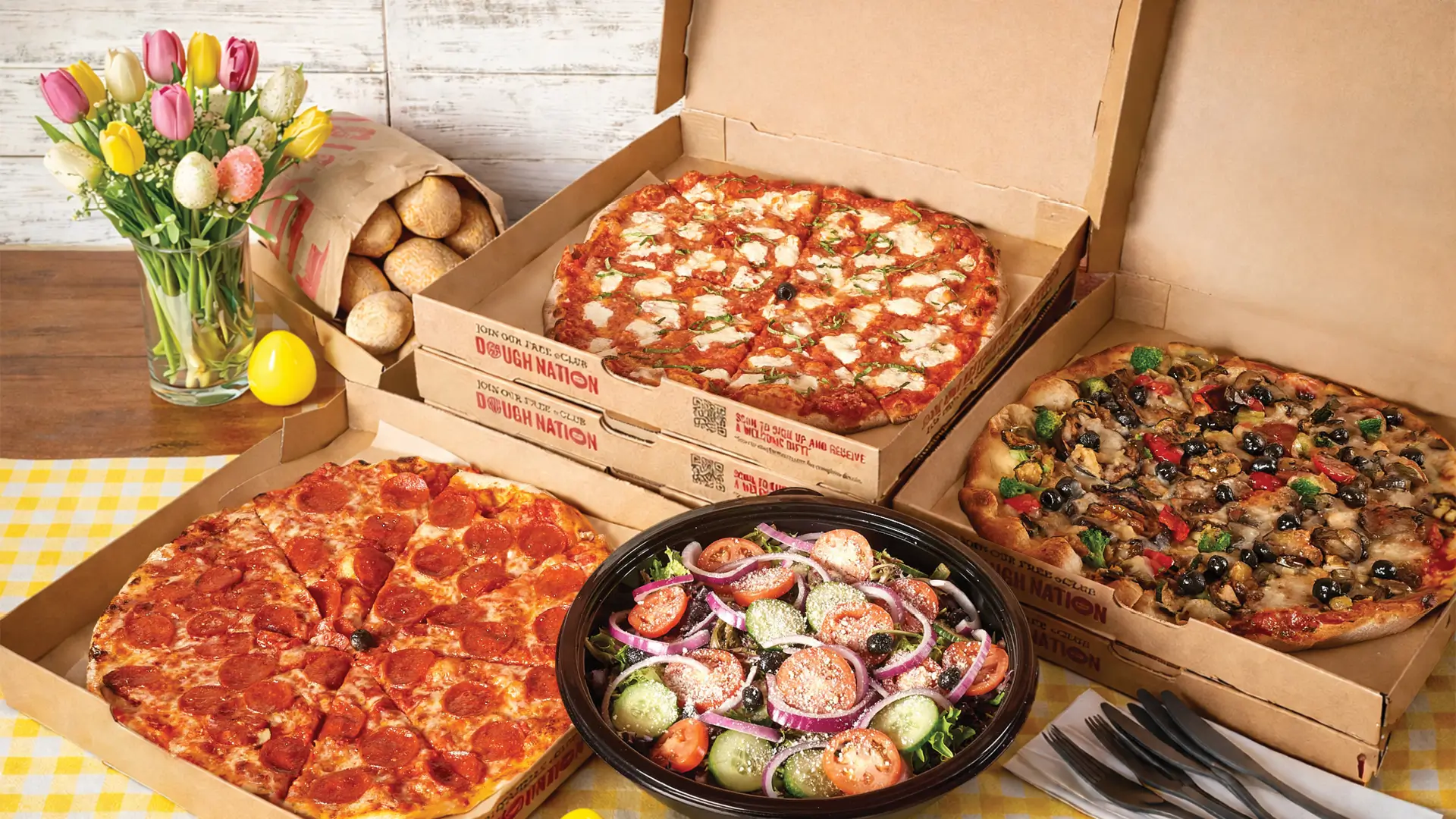Bertucci’s pizzas in open boxes with a fresh garden salad, rolls, and spring flowers with Easter eggs on the table.